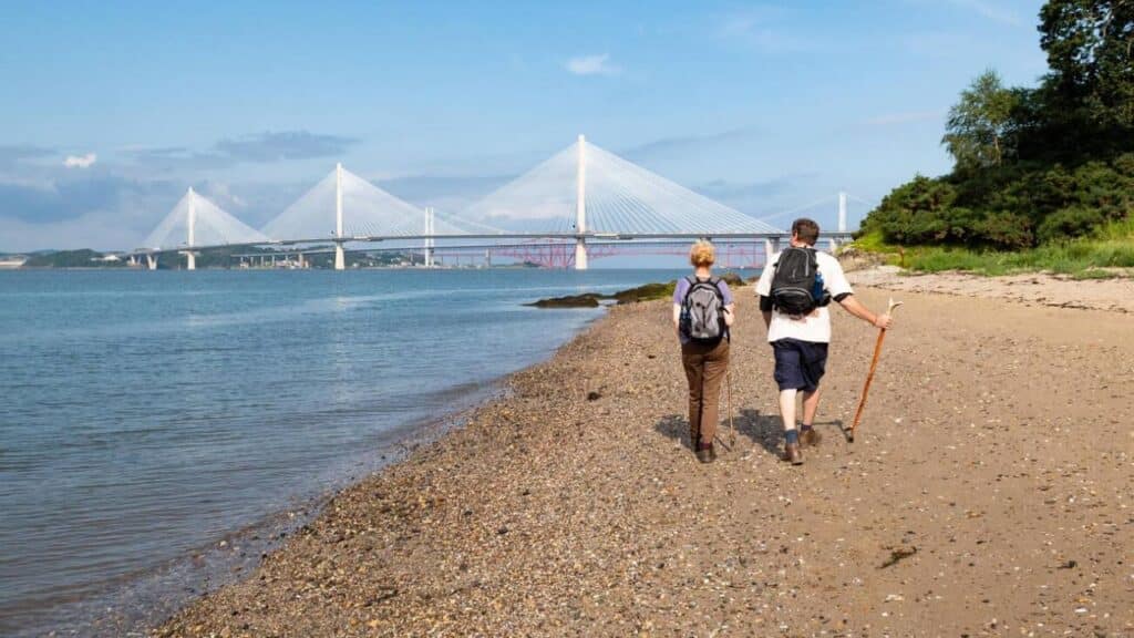 John Muir Way long distance walk - couple walking on beach towards the Queensferry Crossing bridge, Scotland, UK